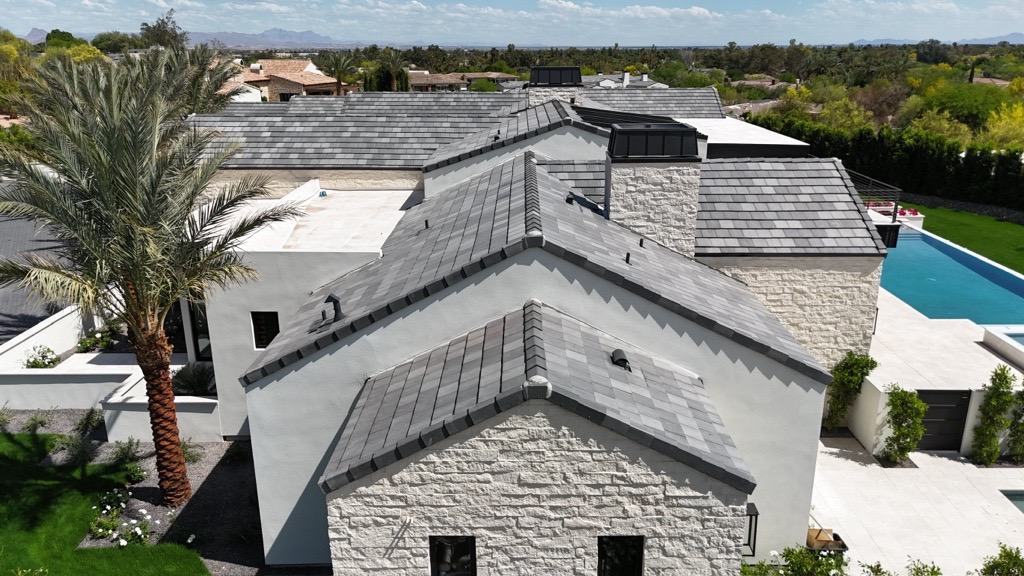 Aerial view of a modern Phoenix home with multiple pitched tile roofs, palm trees, and a backyard pool.
