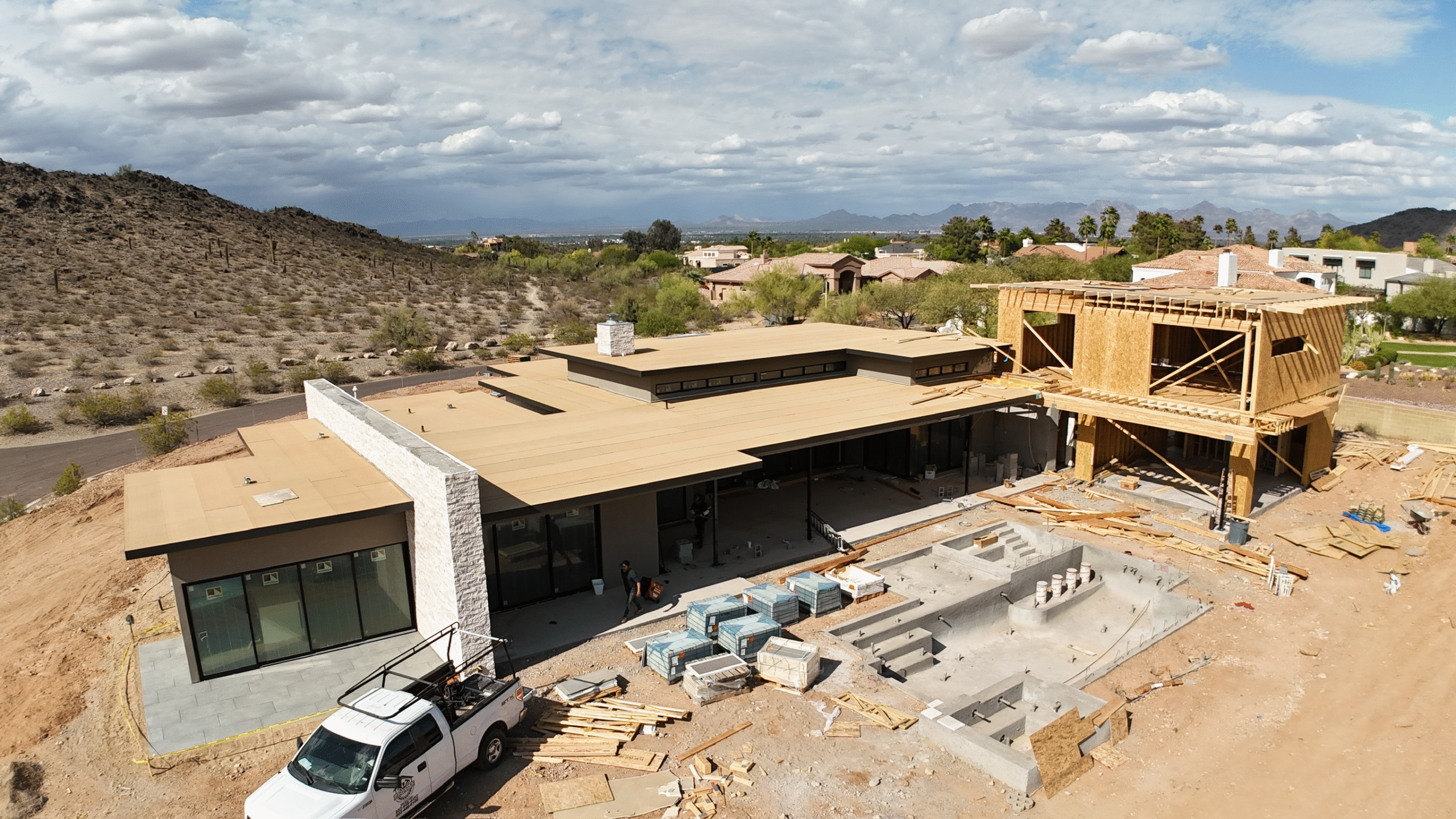 Flat-roof home under construction in Arizona featuring a partially built second story and pool installation, with desert hills and suburban homes in the background.