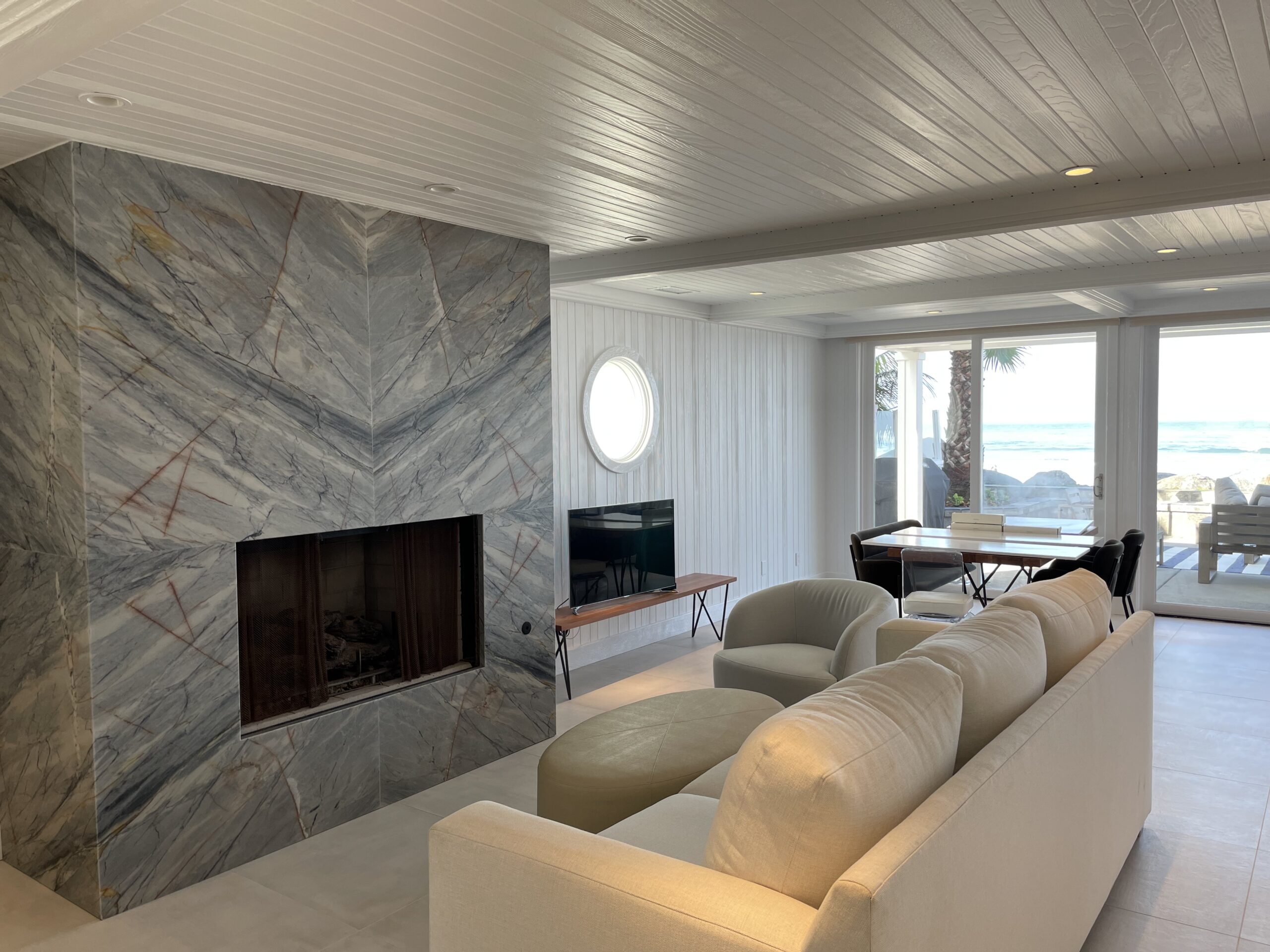 Bright modern living room with white wood-paneled ceiling, marble fireplace, plush cream sofa, and ocean view through large glass doors.