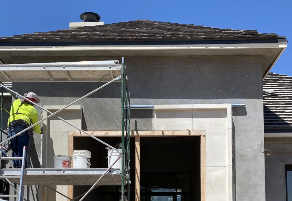 Construction worker on scaffolding applying exterior finish to a modern Arizona home with custom roof and tall entryway.