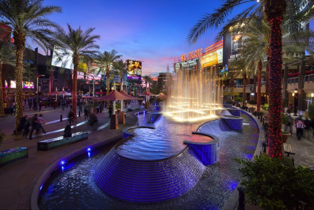 Night view of the vibrant Westgate Entertainment District in Glendale, Arizona, featuring illuminated fountains, palm trees wrapped in lights, and people enjoying the lively atmosphere.